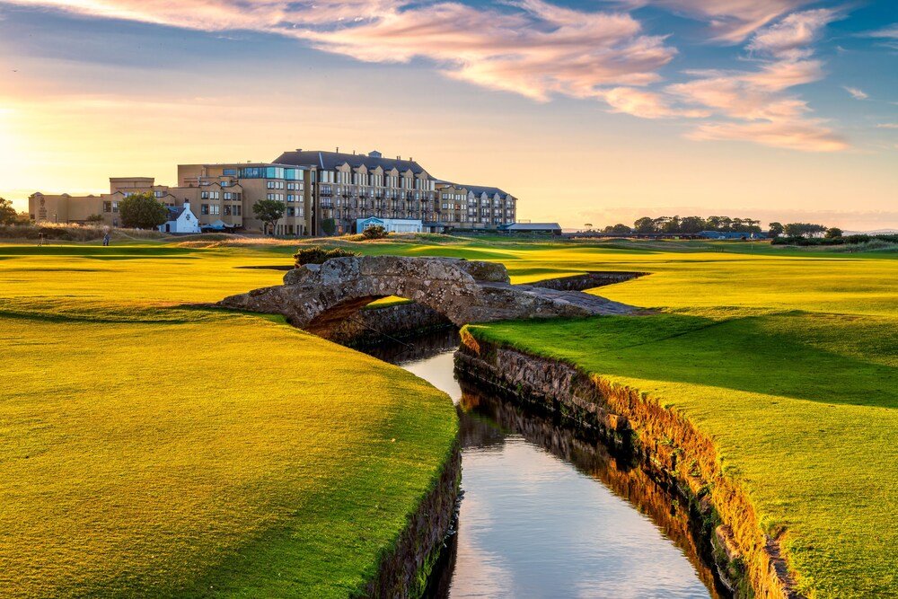 Campo de golf con puente de piedra, gran edificio y cielo al atardecer